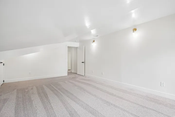 Spacious room with sloped ceilings, light-toned carpet, white walls, recessed ceiling lights, and two wall sconces