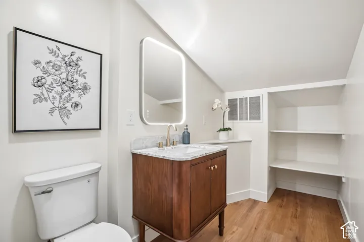 Bathroom featuring a wood-finish vanity with a stone countertop, an illuminated mirror, a white toilet, built-in shelving, and wood-finish flooring
