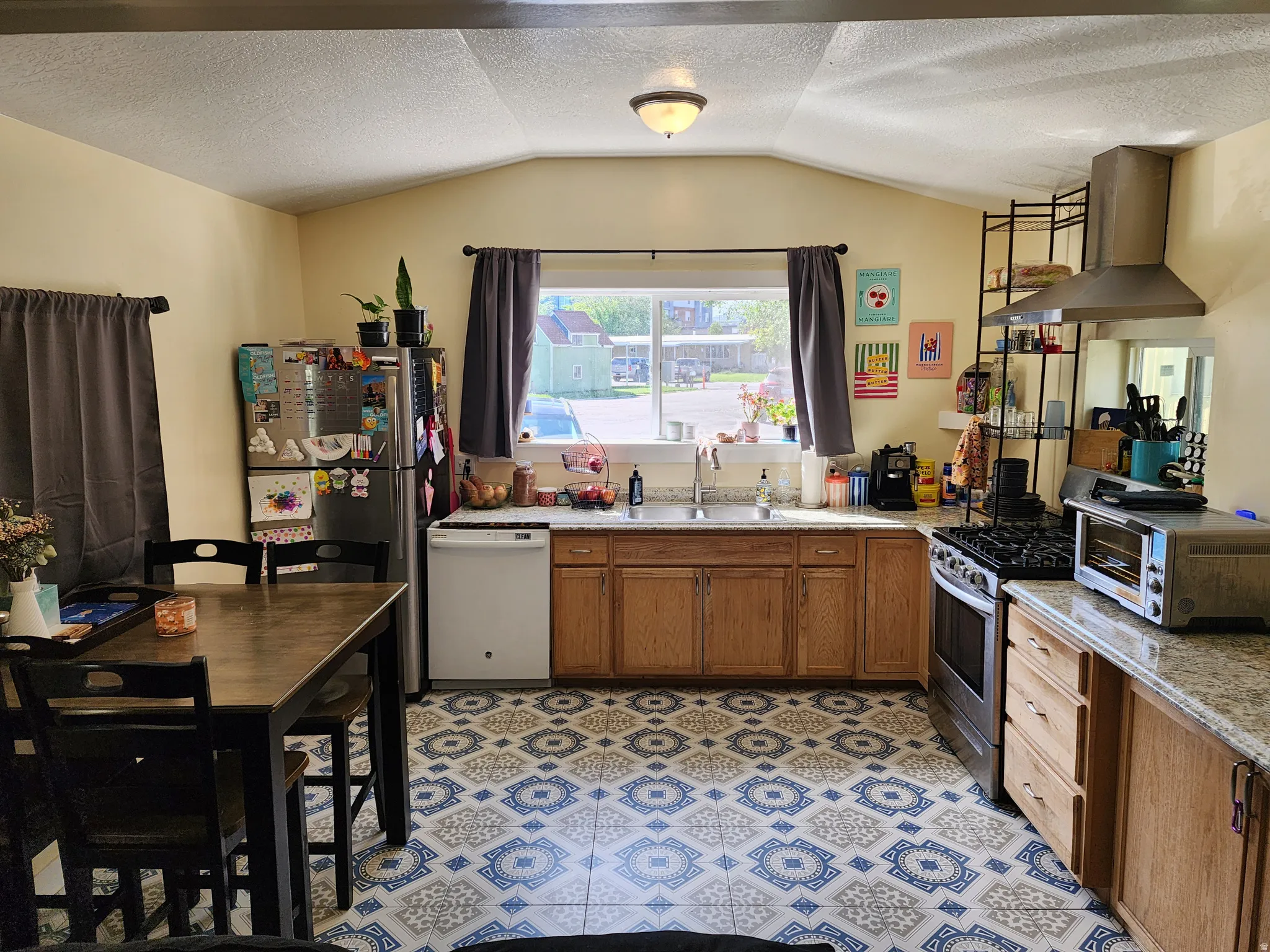 Kitchen featuring patterned floor tiles, light wood-finish cabinetry, granite-style countertops, and a stainless steel gas range with overhead vent hood