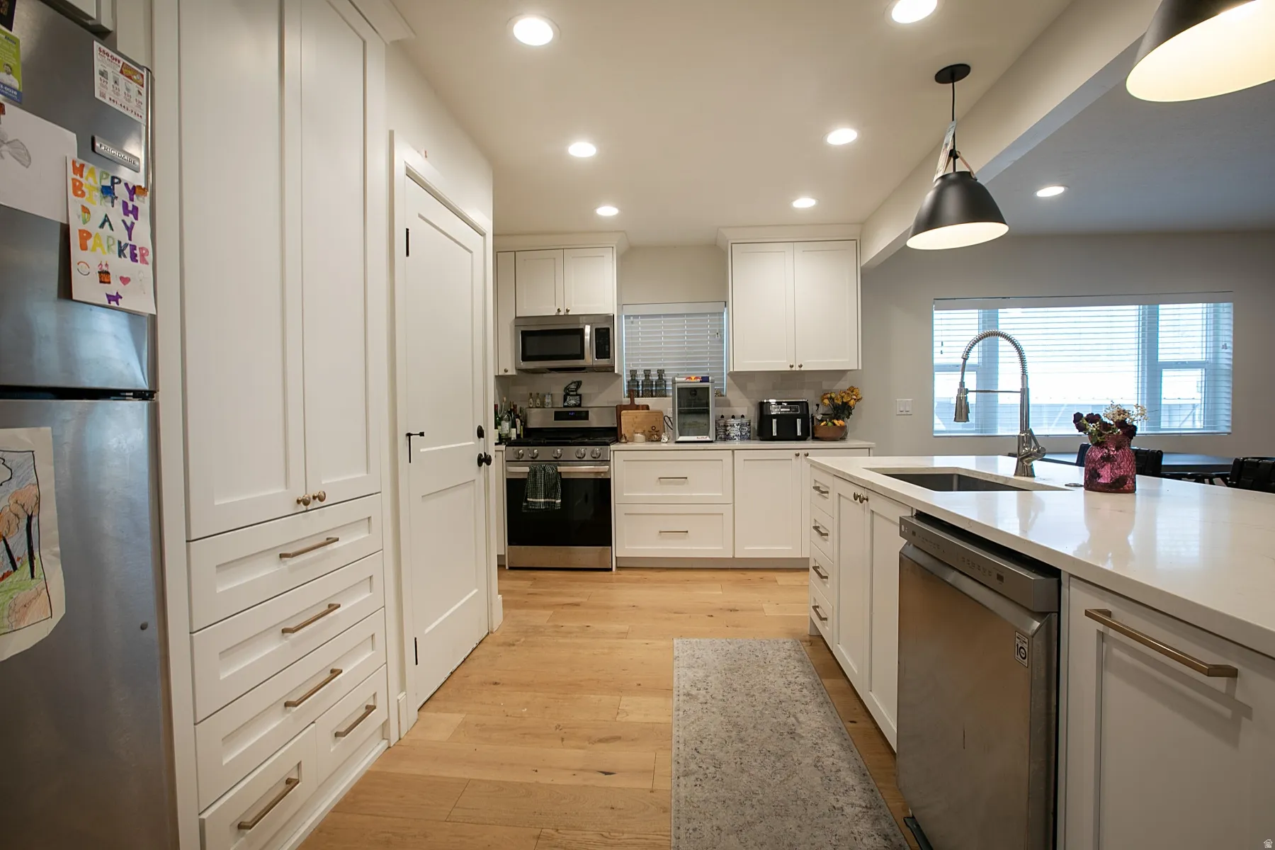 Kitchen with stainless steel appliances, white cabinets, light wood-type flooring, decorative light fixtures, and light stone countertops
