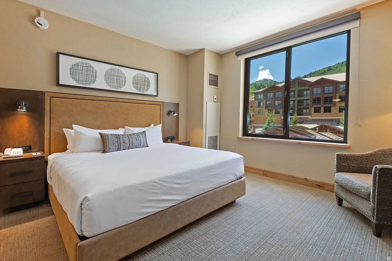 Bedroom featuring a large window with mountain scenery, a neutral color palette, built-in bedside sconces, dark wood-finish nightstands, and textured carpeting