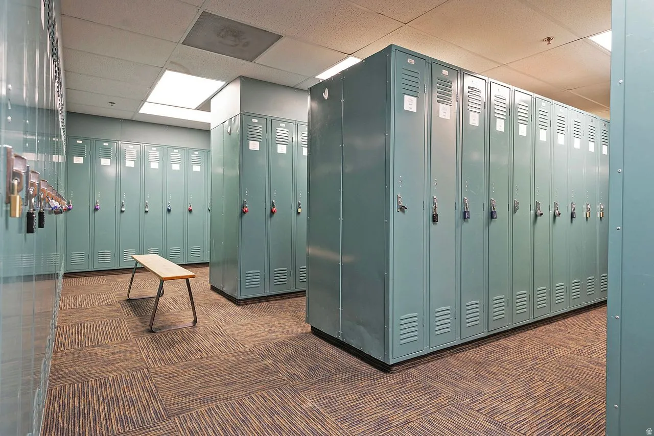 Locker room featuring multiple rows of metal lockers, suspended acoustic ceiling tiles, recessed lighting, and patterned carpet tile flooring