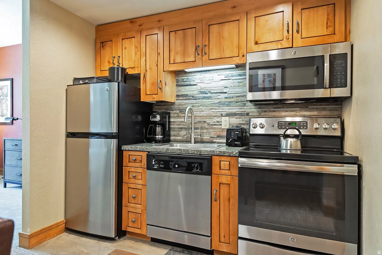 Compact kitchen featuring wood cabinetry, stainless steel appliances, a multi-tone tile backsplash, light-colored countertops, and an undermount sink with a gooseneck faucet