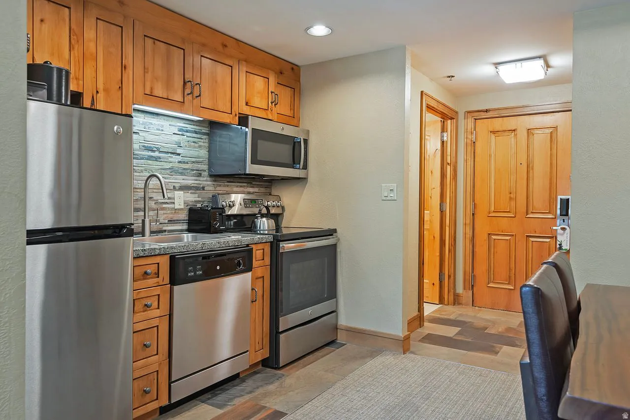 Galley kitchen featuring wood cabinetry, stainless steel appliances, a tile backsplash, and stone-finish countertops