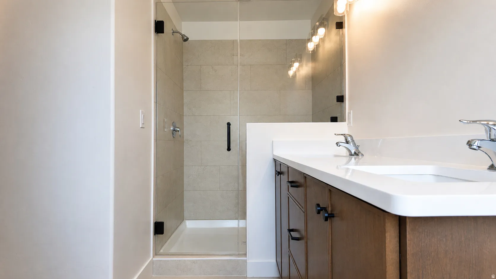 Bathroom featuring a glass-enclosed shower with tile surround, a dual vanity with undermount sinks, white countertops, and wood-finish cabinetry with dark hardware