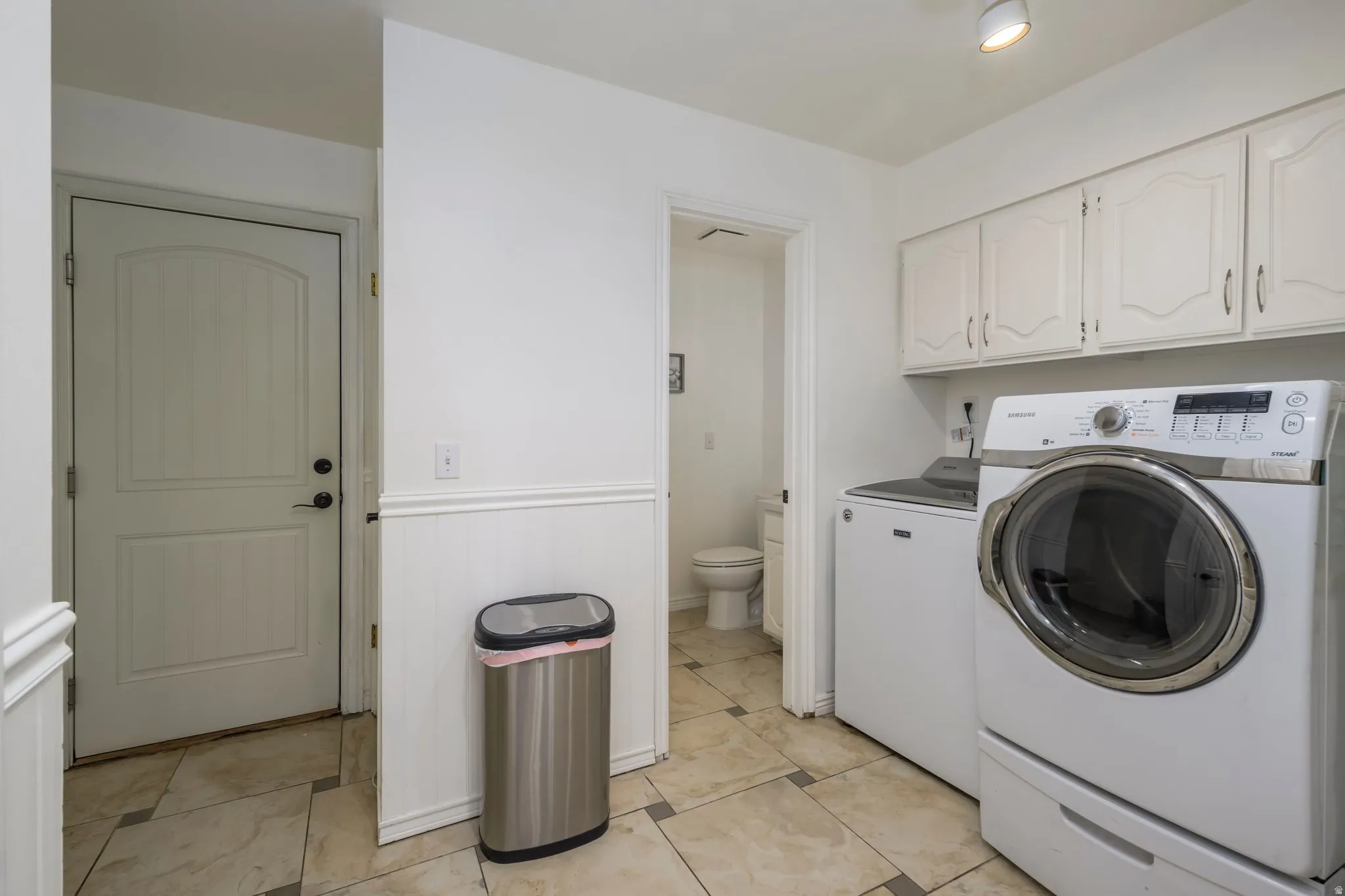 Laundry room featuring tile flooring, white wainscoting, white cabinetry with decorative paneling, and a door with an arched top panel