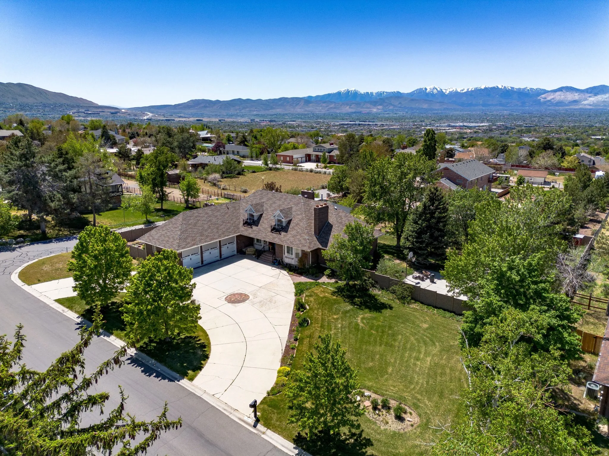 Spacious property featuring a three-car garage, expansive concrete driveway, manicured lawn, mature trees, and a mountain backdrop
