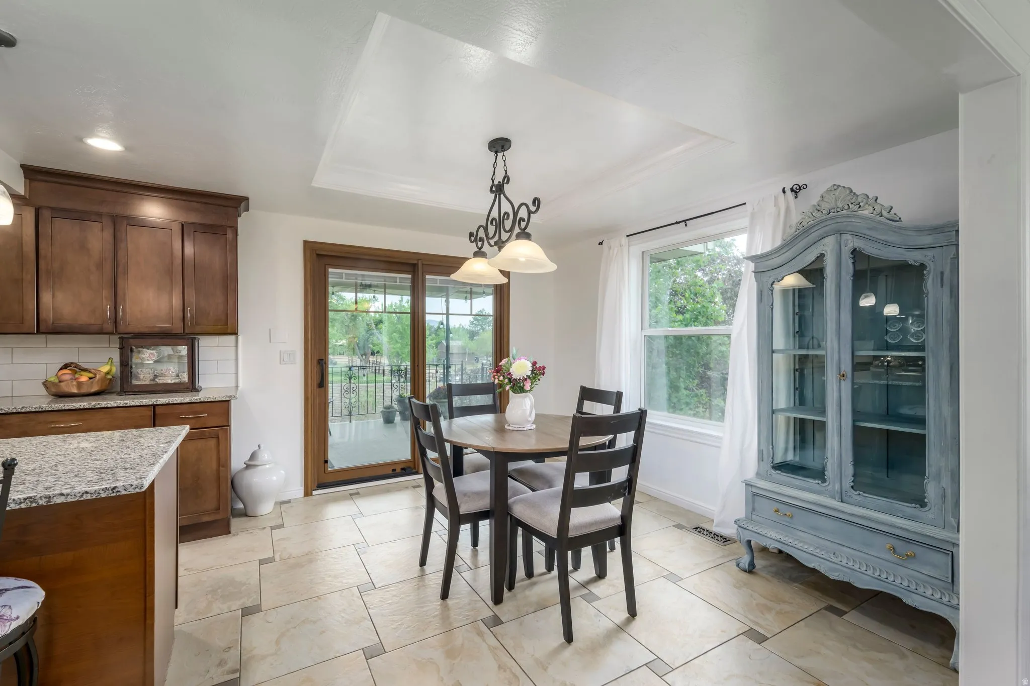 Kitchen and dining area featuring wood cabinetry, light-toned tiled flooring, white subway tile backsplash, a tray ceiling, and a sliding glass door