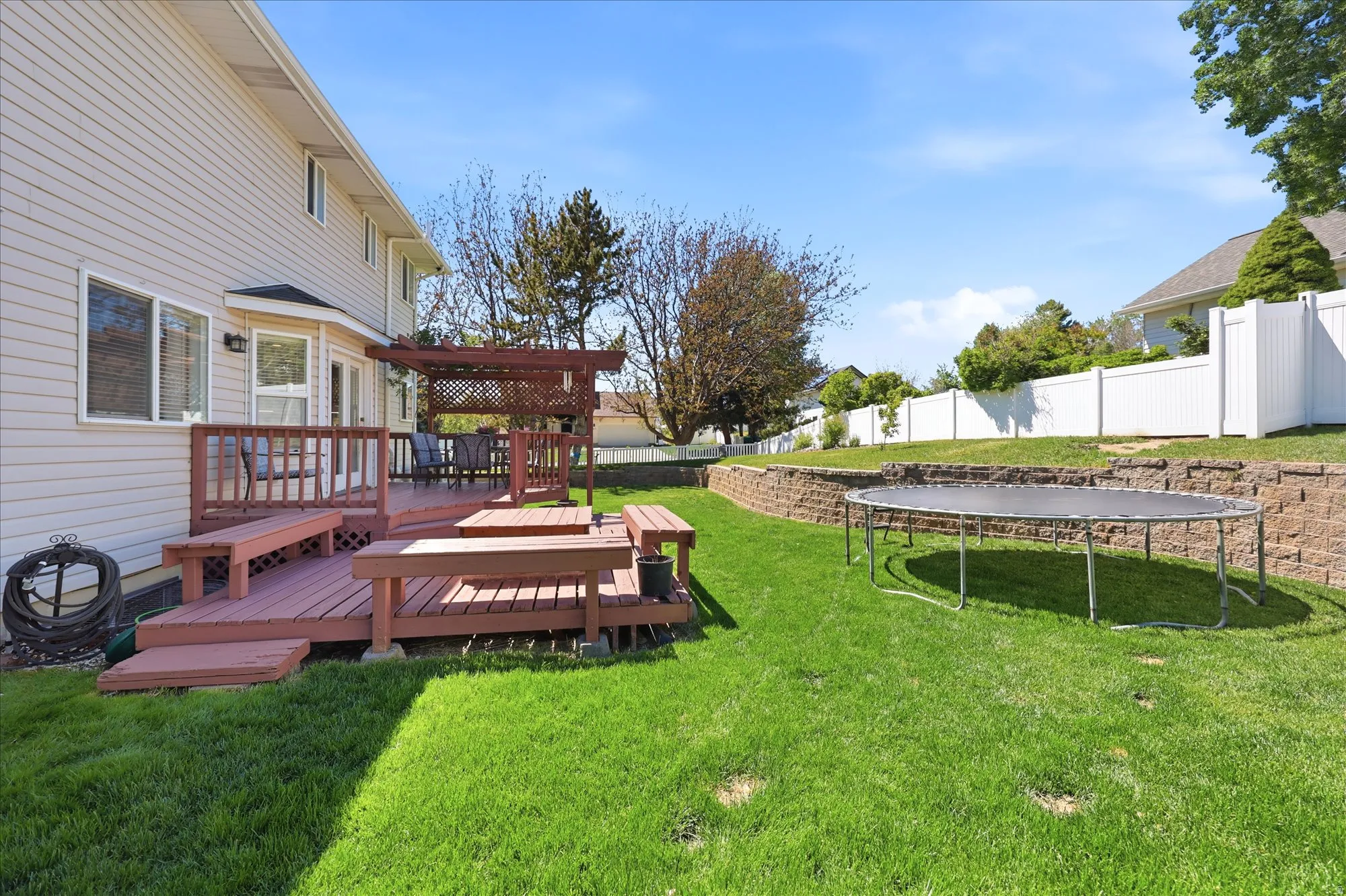 Expansive backyard featuring a multi-level wood deck with built-in benches, a decorative pergola, a manicured lawn, a retaining wall, and a white privacy fence