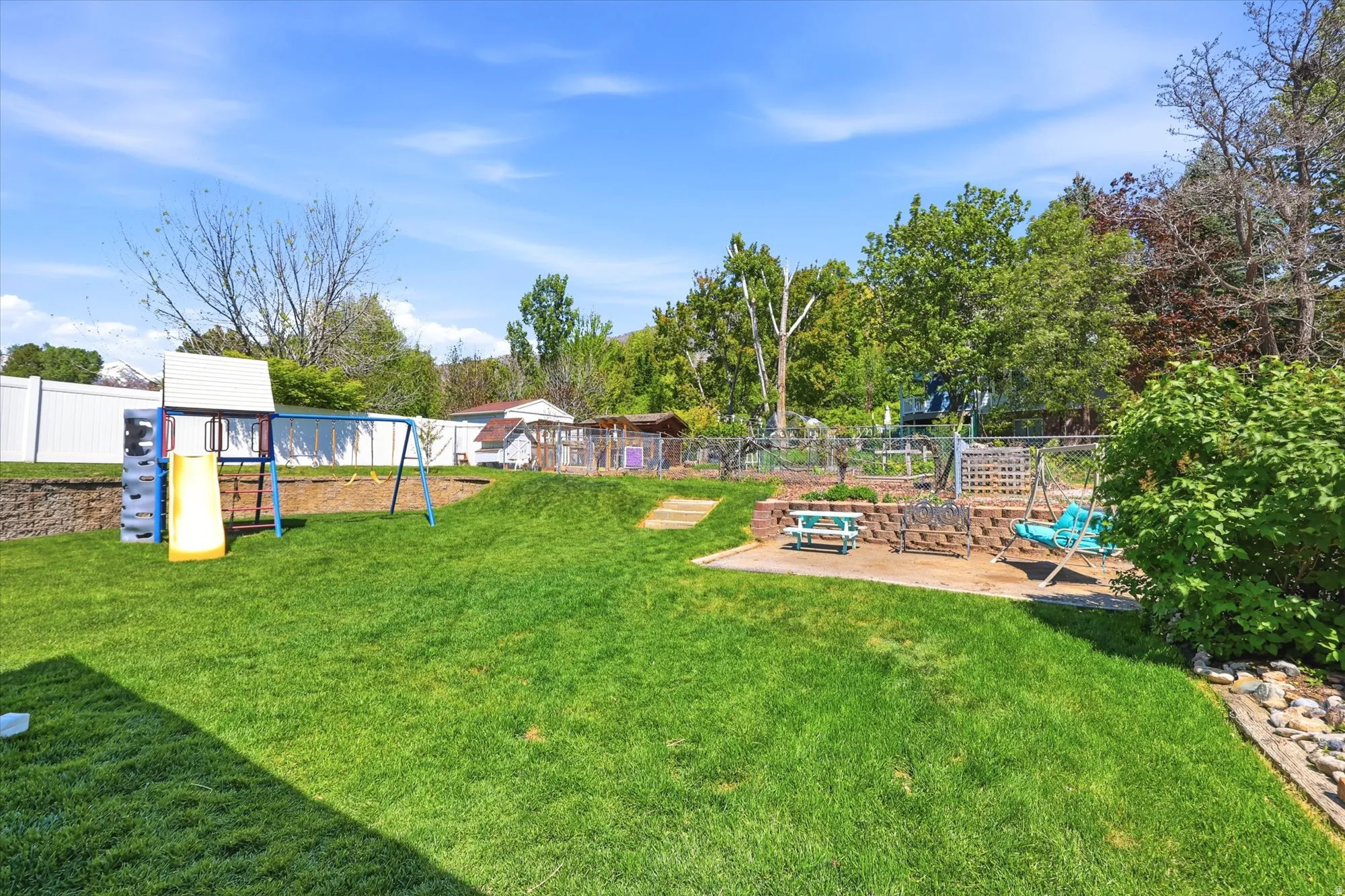 Expansive backyard featuring a tiered landscape, a manicured lawn, a white privacy fence, mature trees, and a paved patio area