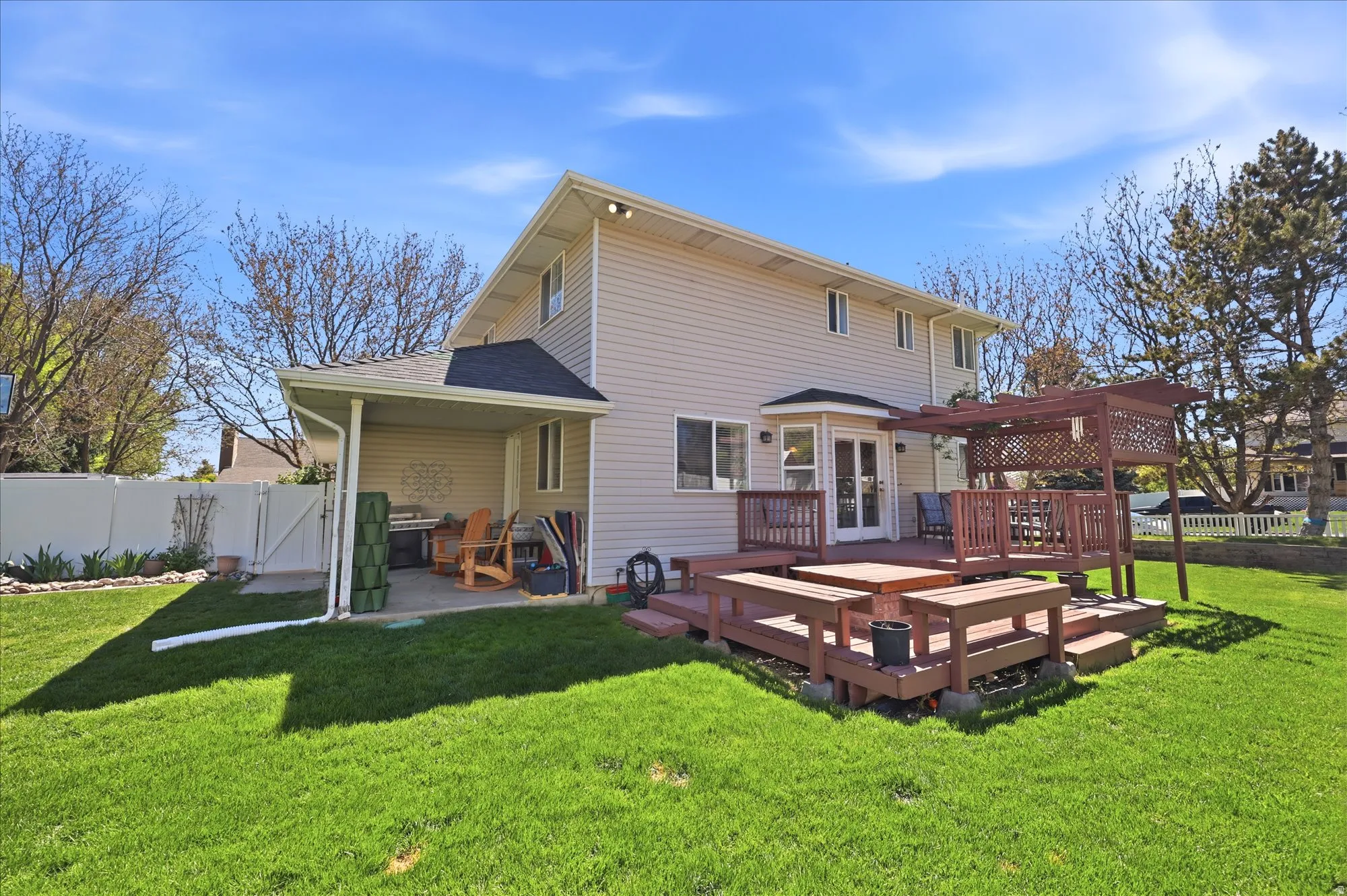 Expansive backyard featuring a multi-level wooden deck with integrated seating, a wooden pergola, a covered concrete patio, and a white privacy fence