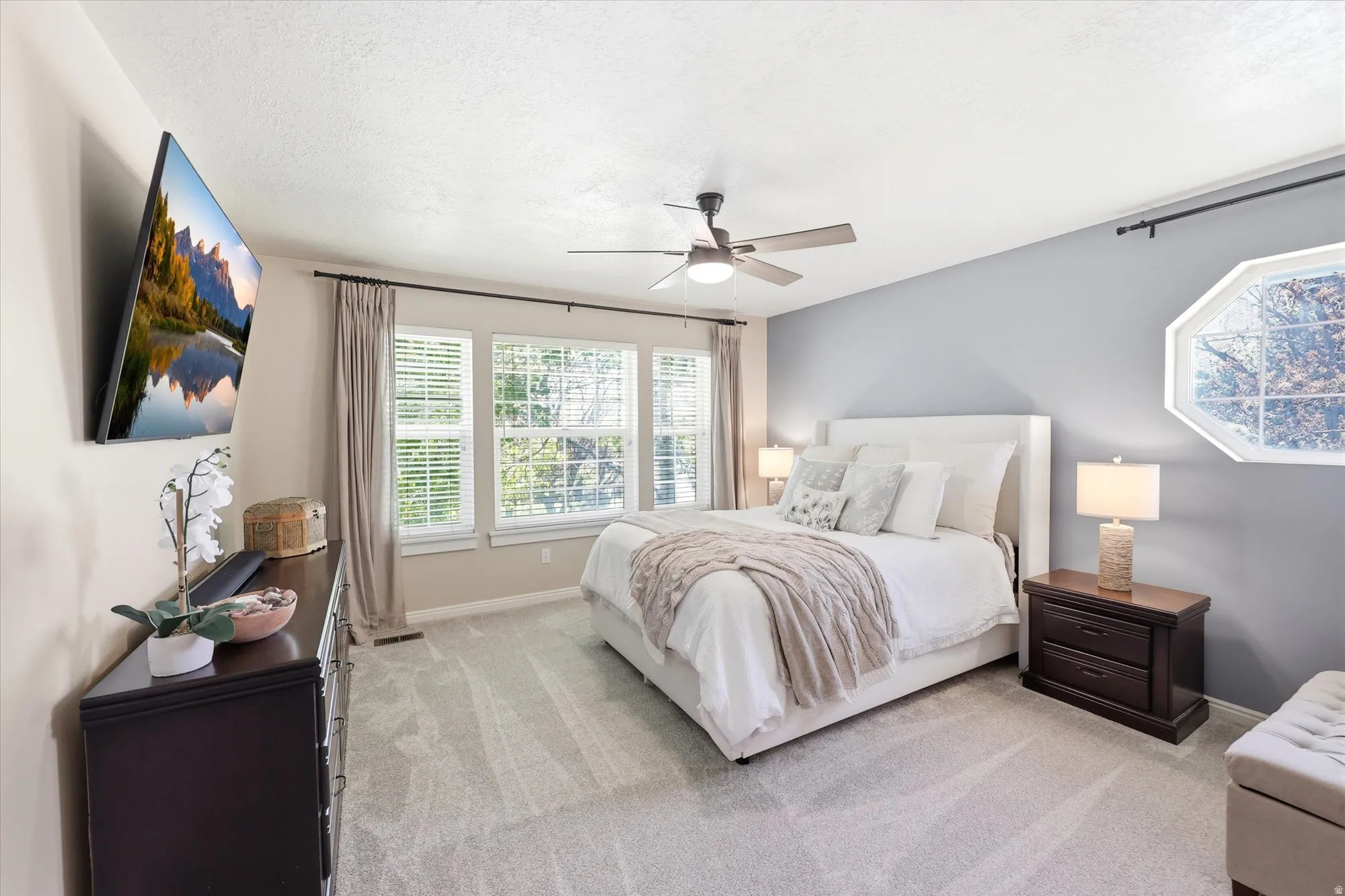 Carpeted bedroom featuring a ceiling fan, multiple windows with blinds, and a built-in wall-mounted television