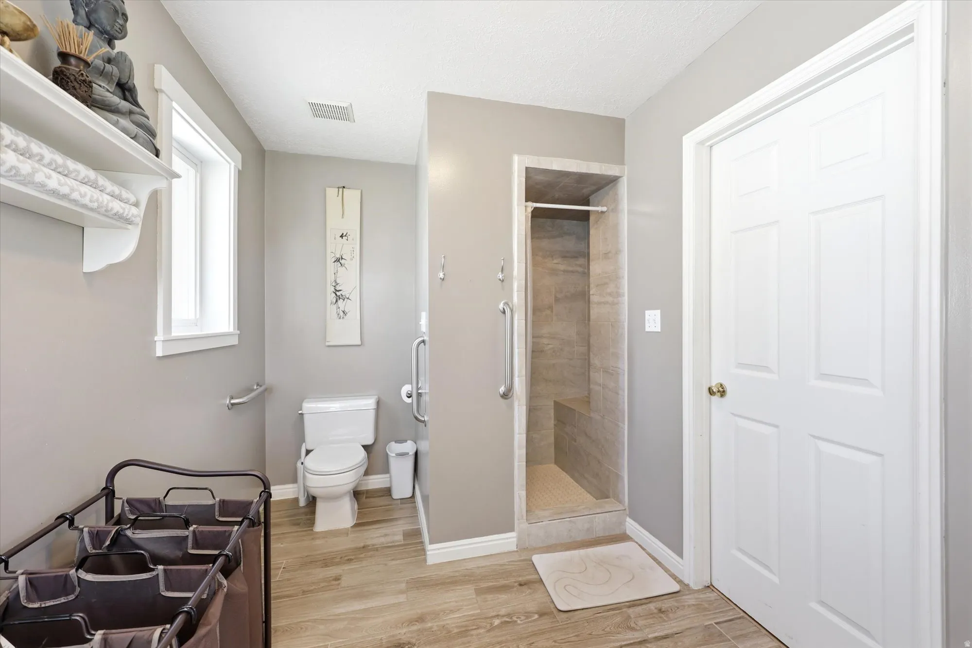 Bathroom featuring wood-finish flooring, a toilet, and an accessible shower with a tiled bench and grab bar