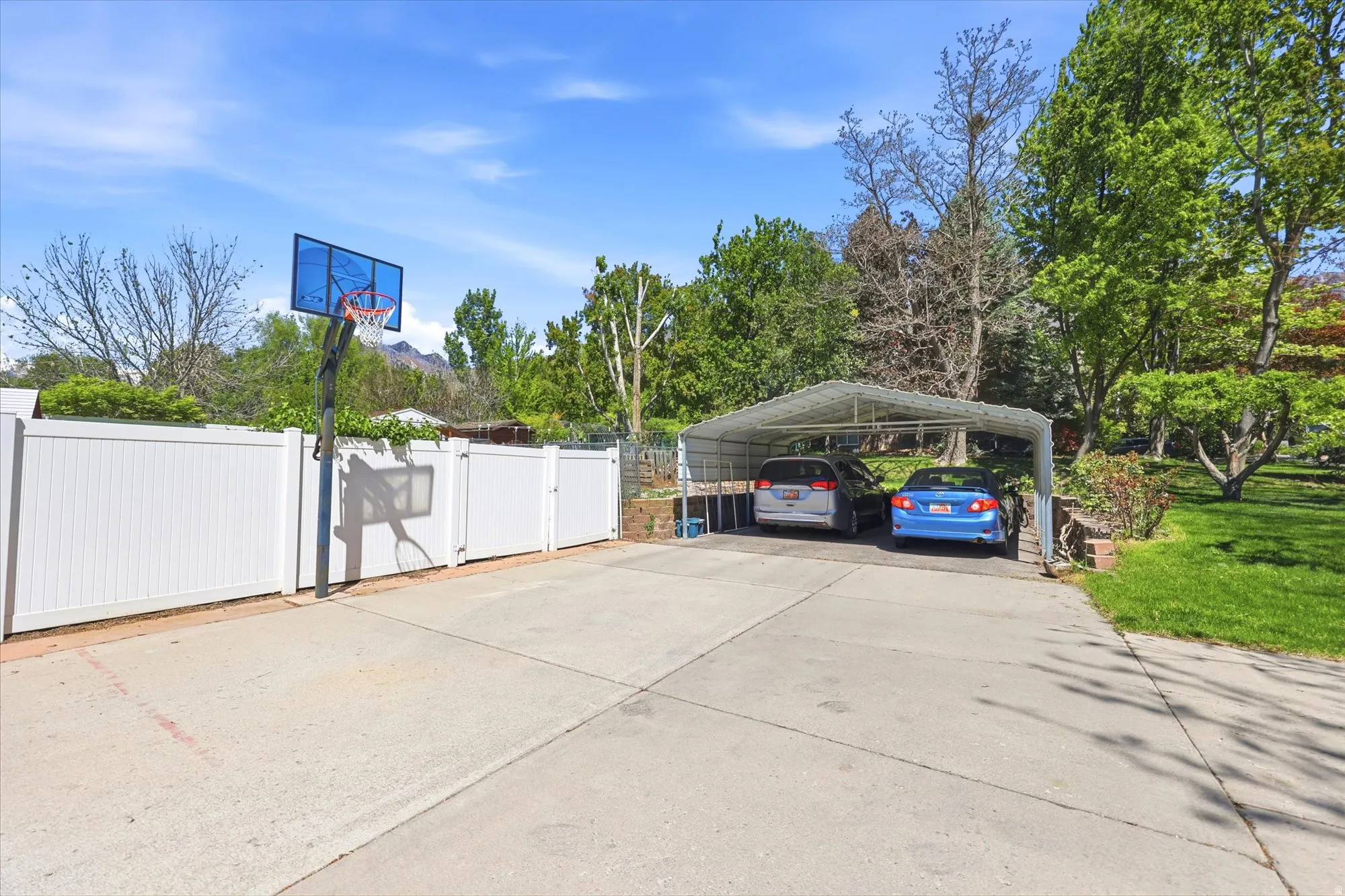 Concrete pad featuring a basketball hoop, white panel fencing, and a metal carport