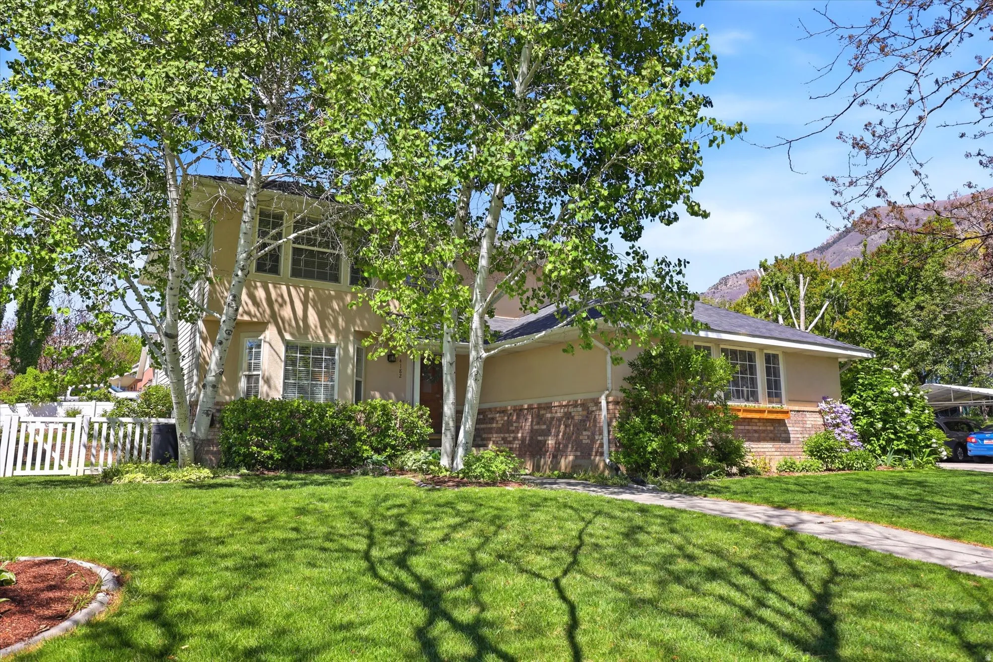 Two-story stucco and brick-clad residence with a pitched roof, front-facing gables, mature trees, a manicured lawn, and a white picket fence