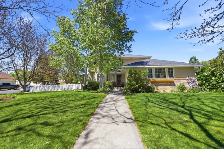 Front exterior featuring a concrete walkway, expansive lawn, mature deciduous trees, a white picket fence, and a multi-level roofline