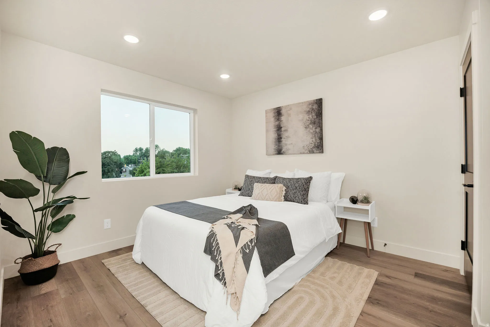 Bright secondary bedroom featuring recessed ceiling lighting, large windows with natural light, wood-finish flooring, and a dedicated nightstand with integrated storage