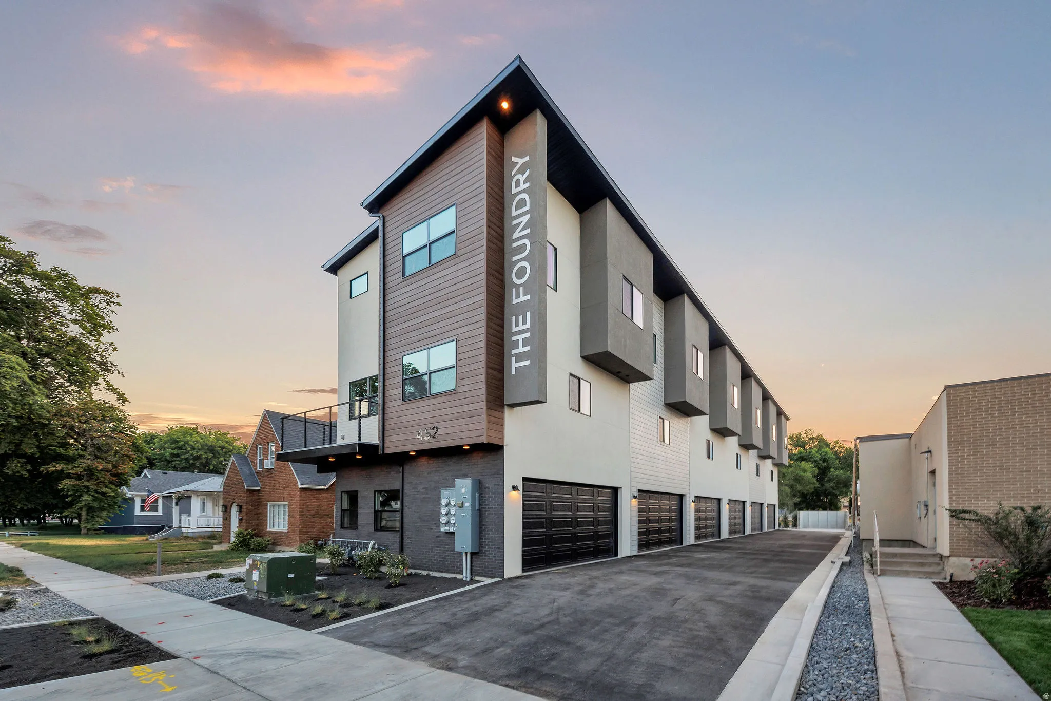 Modern multi-story building featuring a wood-finish facade, contemporary architectural elements, individual garages, clean landscaping, and a concrete sidewalk