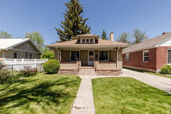Brick facade residence with a covered porch, stone foundation accents, a concrete walkway, a paved driveway, and a front lawn
