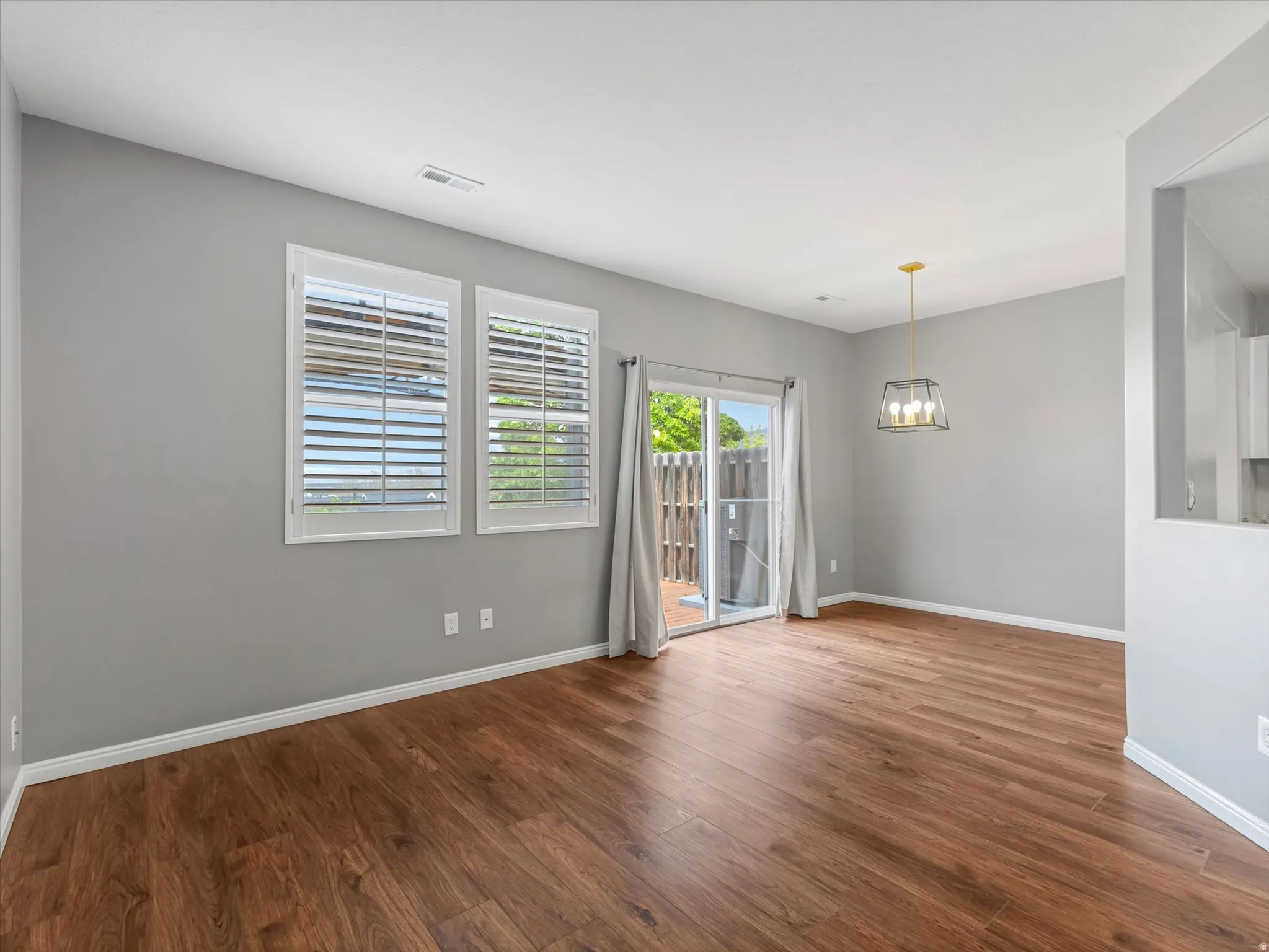 Spacious living area featuring wood-finish flooring, two windows with plantation shutters, a sliding glass door, and a modern geometric pendant light fixture