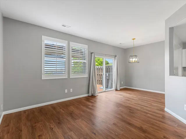Spacious living area featuring wood-finish flooring, two windows with plantation shutters, a sliding glass door, and a modern geometric pendant light fixture