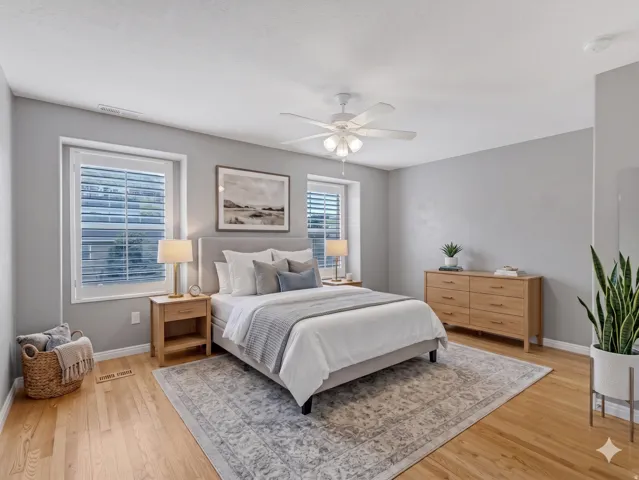 Bright bedroom featuring wood-finish flooring, two large windows with blinds, a ceiling fan with integrated lighting, and light gray wall tones