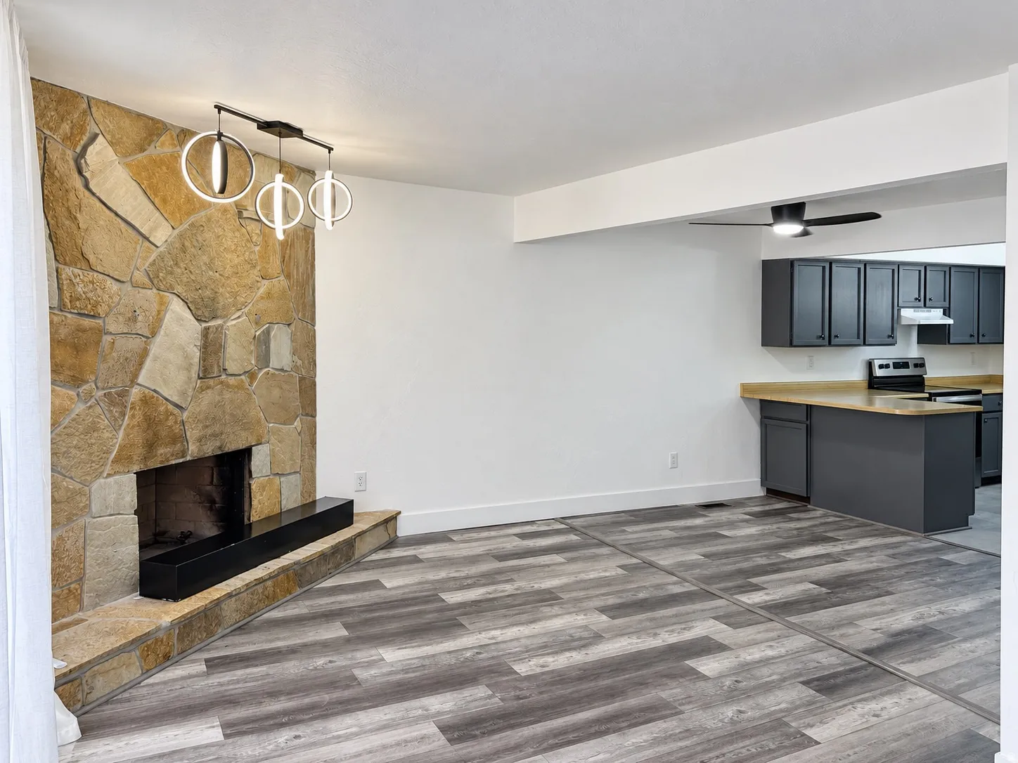 Stone-clad fireplace with a hearth, modern chandelier, wood-finish flooring, and a kitchen with dark cabinetry and light countertops