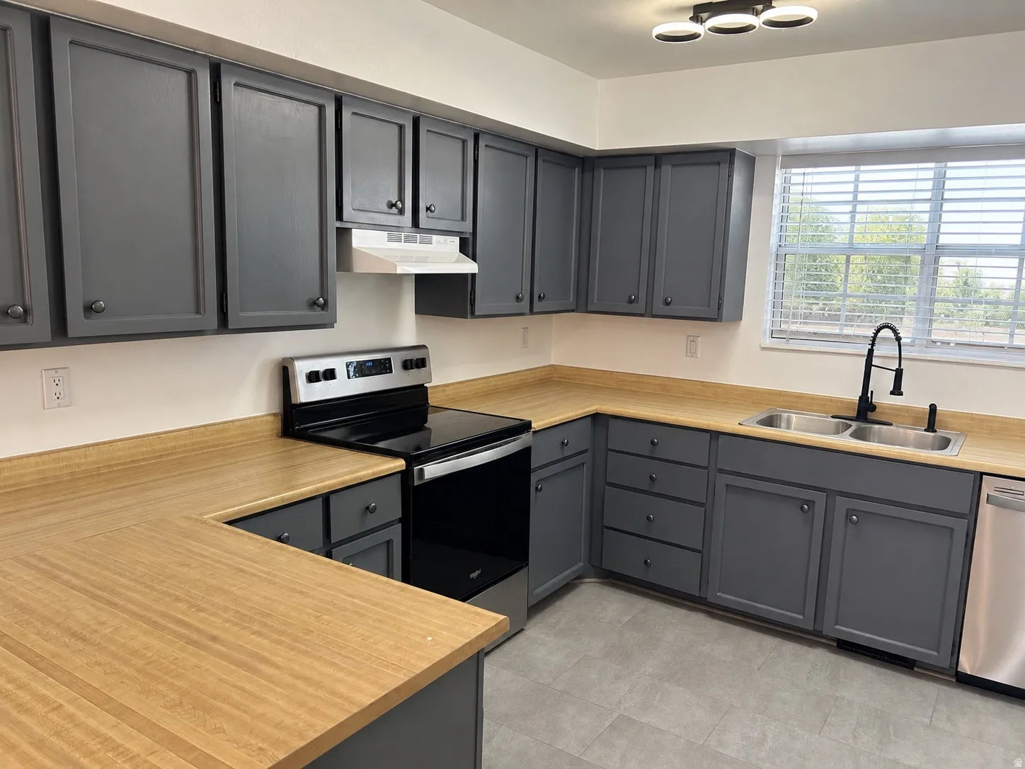 Kitchen featuring gray cabinetry, light wood-finish countertops, stainless steel appliances, a double basin sink with a matte black faucet, and a contemporary ceiling light fixture