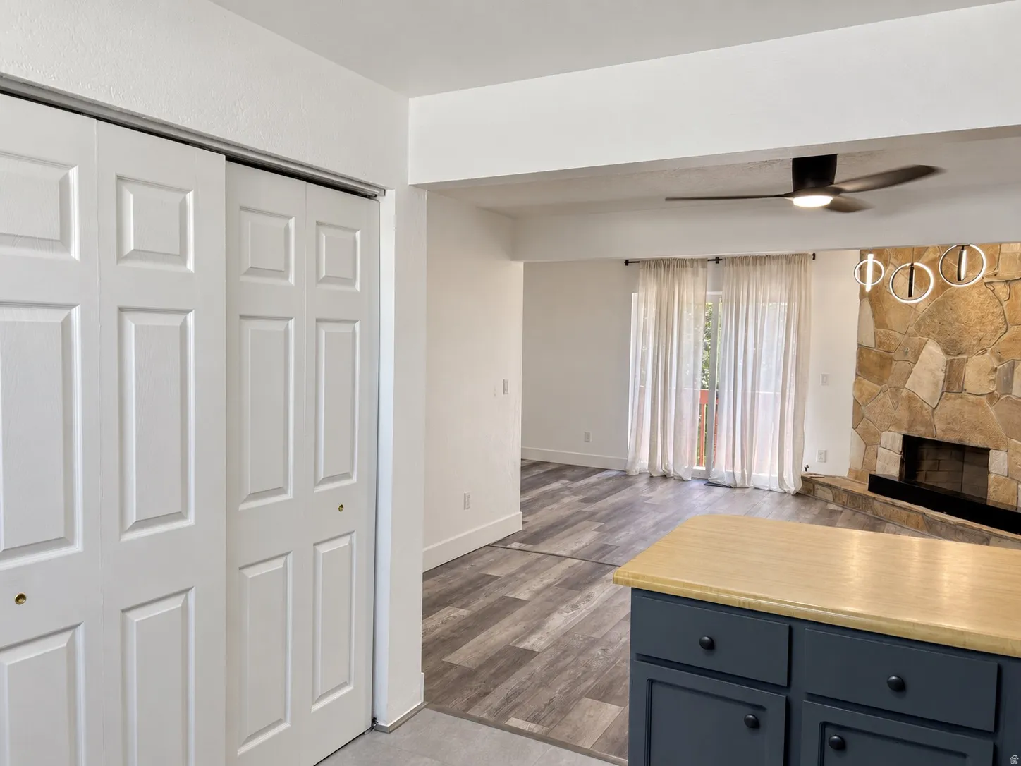 Built-in storage with bi-fold doors, kitchen island with wood-finish countertop and dark cabinetry, wood-finish flooring, stone fireplace, and ceiling fan with integrated lighting