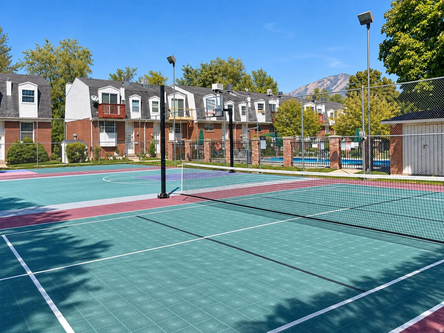 Multi-sport court with a tennis net, adjacent to a pool area with fencing, and brick-exterior residential buildings
