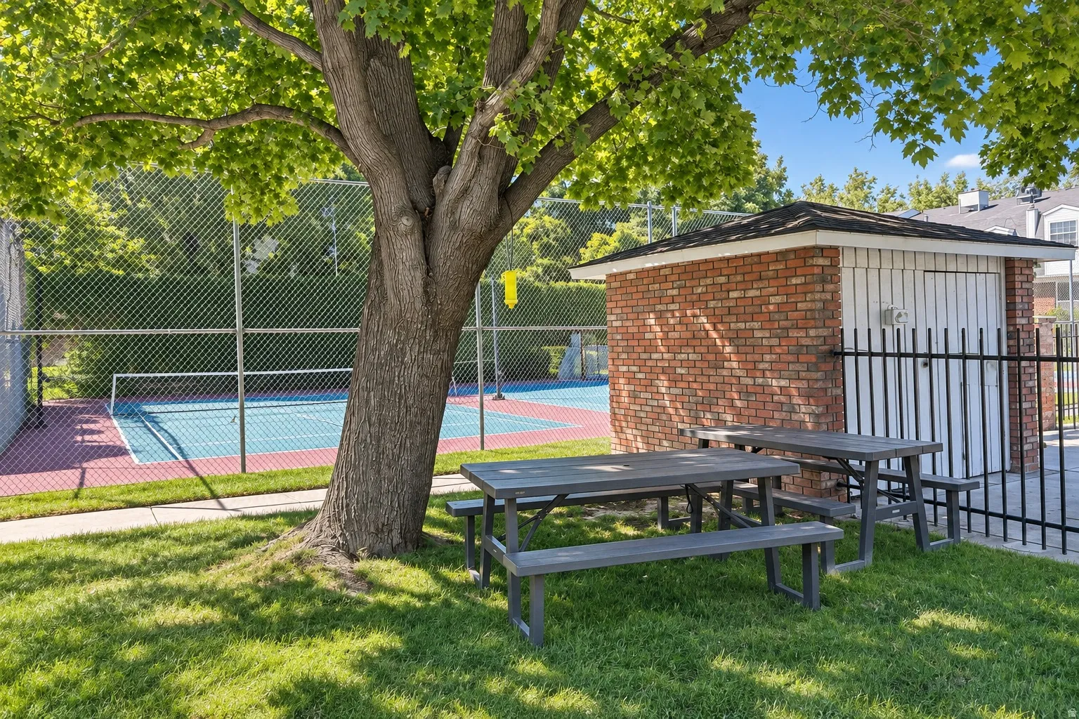 Recreational area featuring a tennis/pickleball court, chain-link fencing, mature trees, and a brick storage shed with a shingled roof