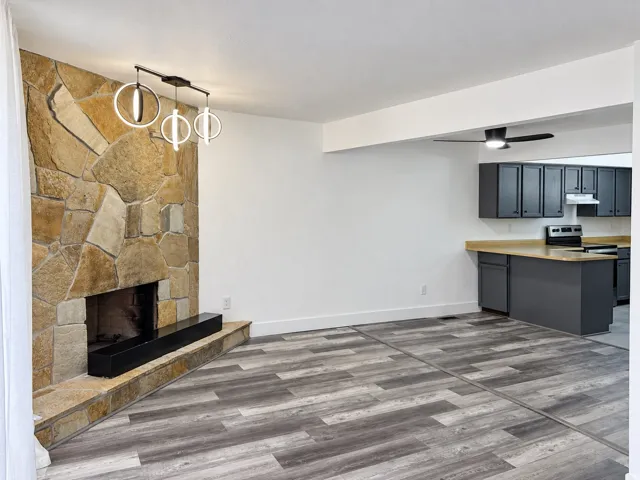 Stone-clad fireplace with a hearth, modern chandelier, wood-finish flooring, and a kitchen with dark cabinetry and light countertops