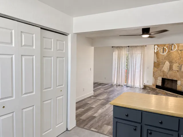 Built-in storage with bi-fold doors, kitchen island with wood-finish countertop and dark cabinetry, wood-finish flooring, stone fireplace, and ceiling fan with integrated lighting