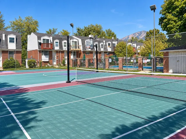 Multi-sport court with a tennis net, adjacent to a pool area with fencing, and brick-exterior residential buildings