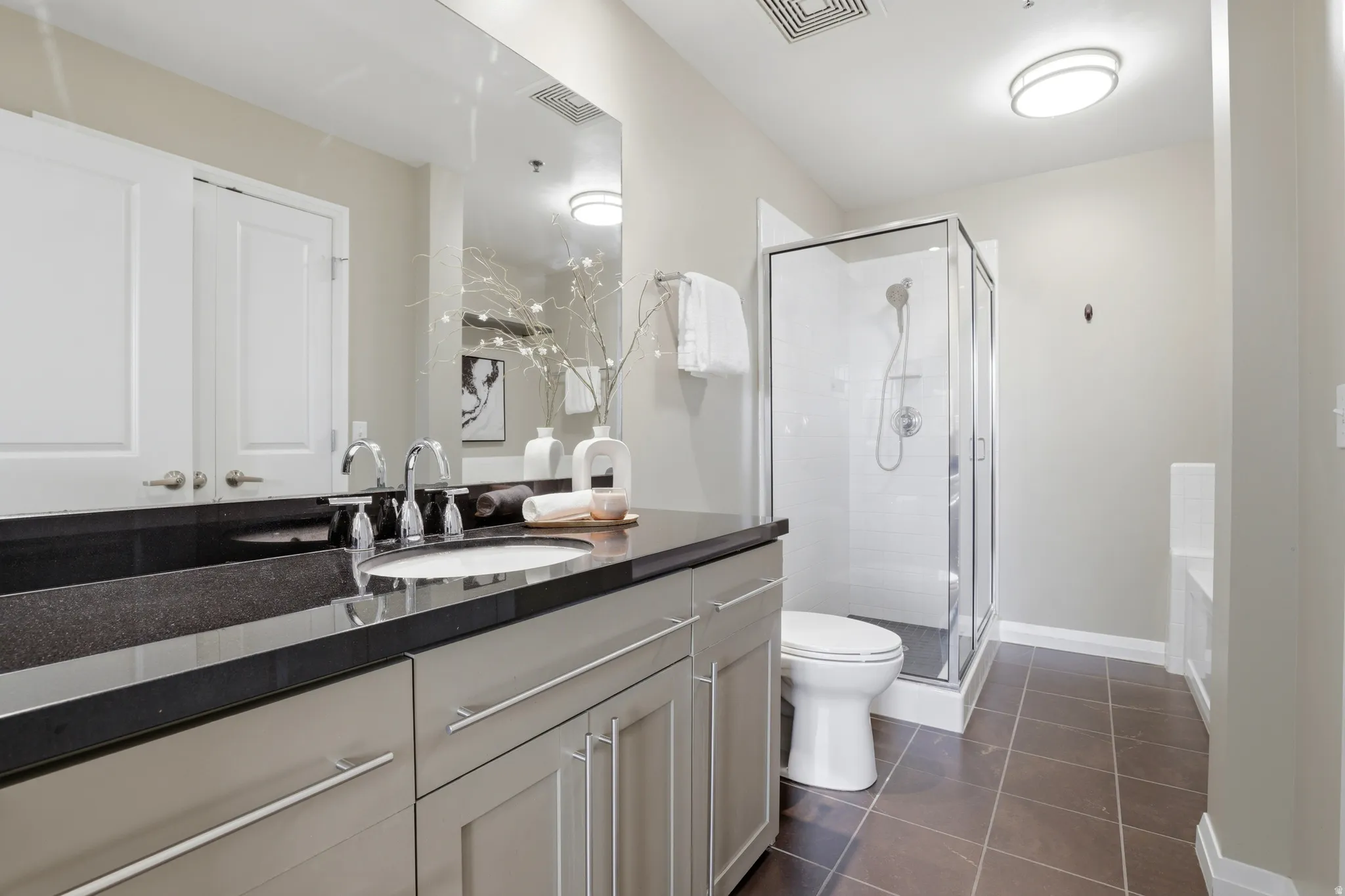 Bathroom featuring a single vanity with a dark countertop and undermount sink, a framed glass shower enclosure, a white toilet, and brown tile flooring