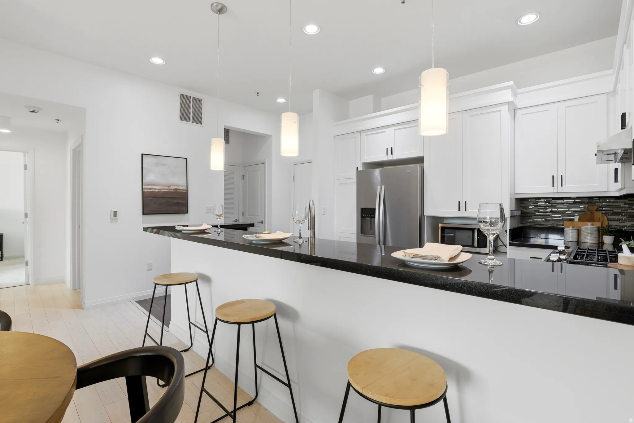 Contemporary kitchen featuring a breakfast bar with dark countertops, white cabinetry, stainless steel appliances, a stacked stone backsplash, and recessed lighting