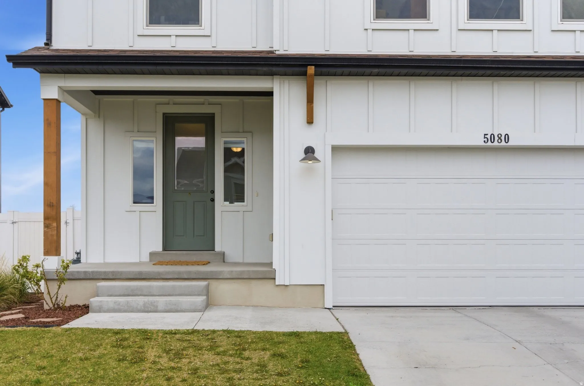 Contemporary white board and batten exterior with a dark green entry door, covered porch featuring a wood accent pillar, integrated garage, and a modern outdoor wall sconce