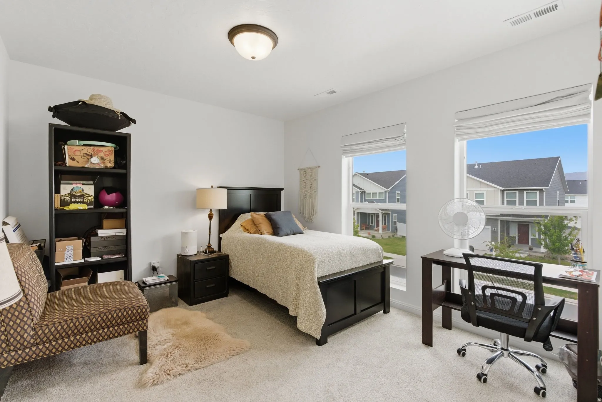 Bright bedroom featuring light gray carpeting, two large windows with Roman shades, a dark wood-finish bed frame, and a flush-mount ceiling light