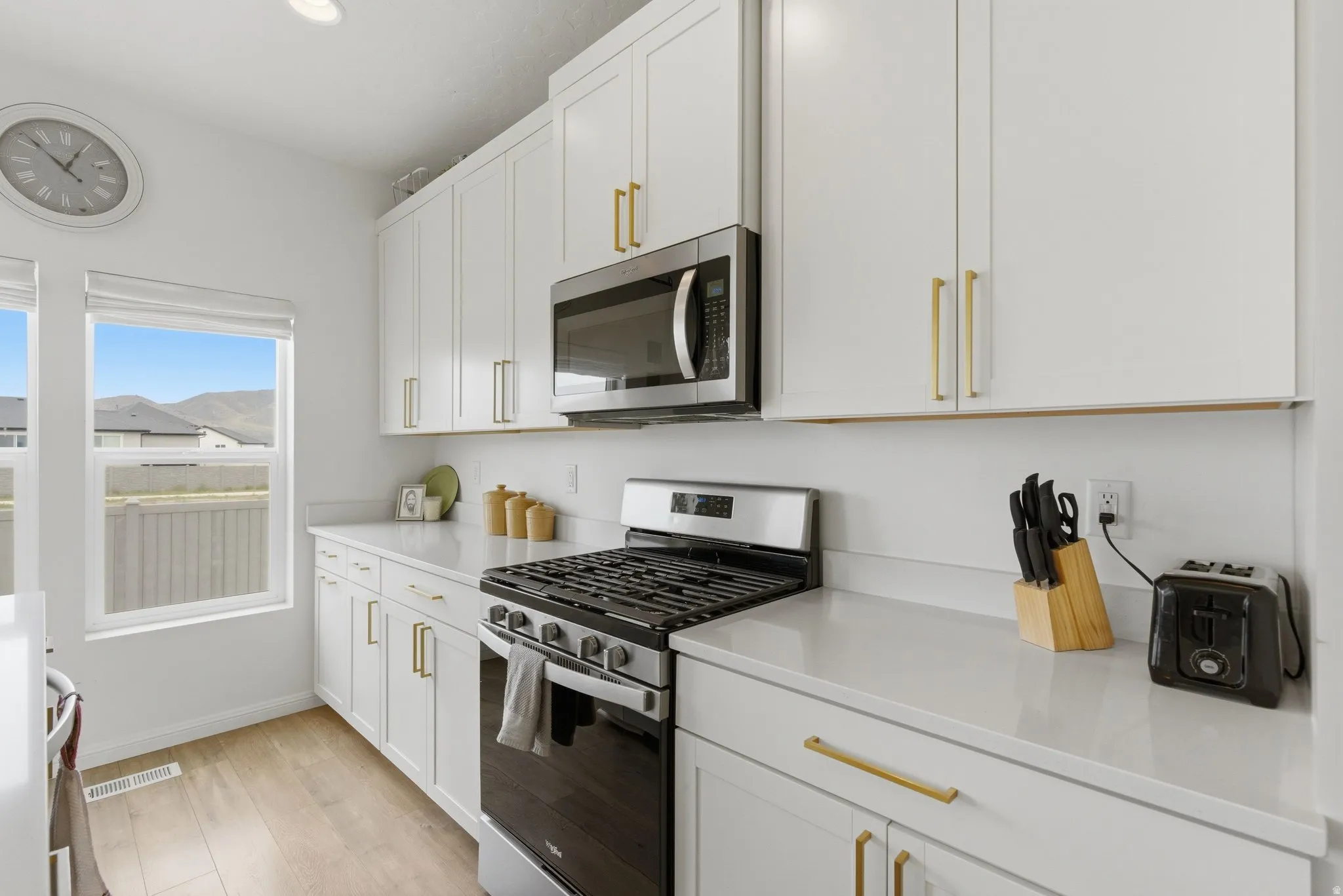 Kitchen featuring white shaker-style cabinetry, gold-tone hardware, stainless steel appliances, light-toned countertops, and wood-finish flooring