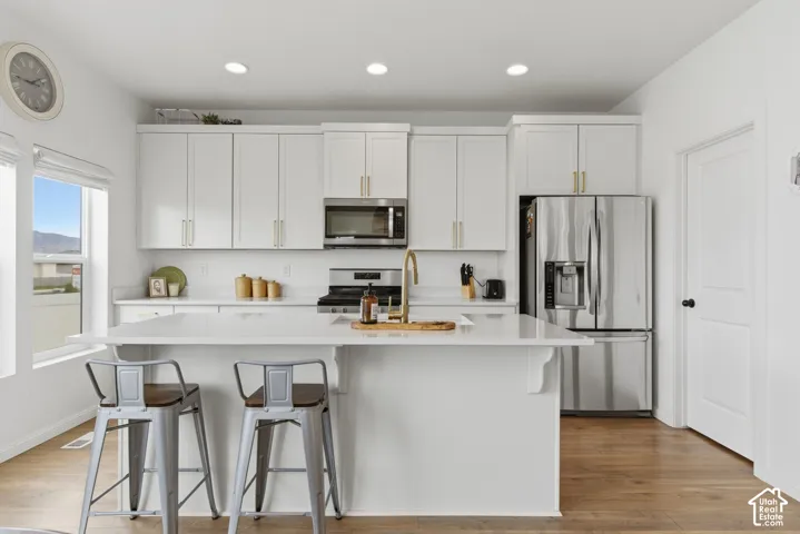 Modern kitchen featuring a central island with countertop seating, white cabinetry with gold-tone hardware, stainless steel appliances, recessed lighting, and wood-finish flooring