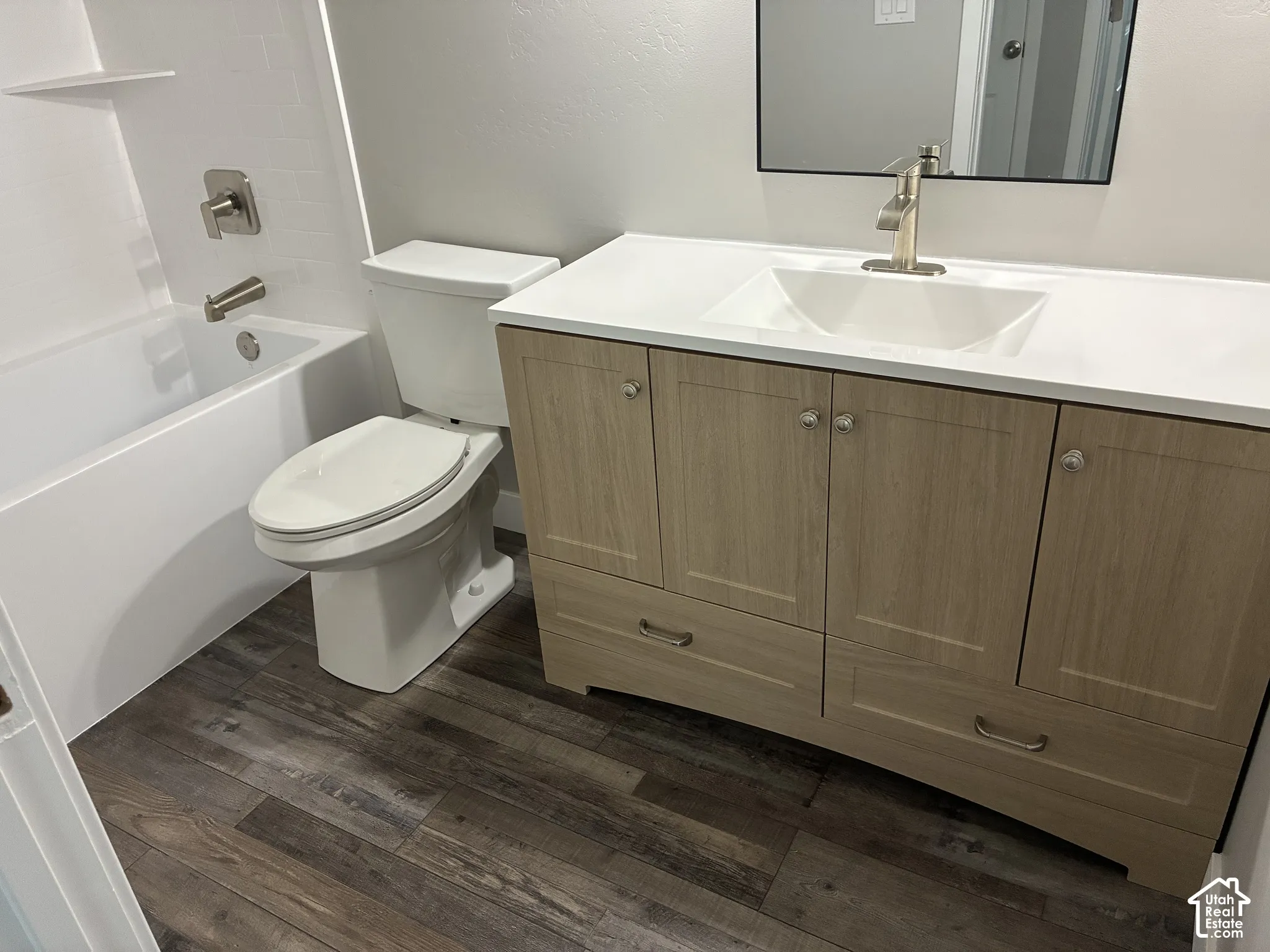 Wood-finish flooring, white bathtub with brushed nickel fixtures, contemporary vanity with a white countertop, under-mount sink, and framed mirror