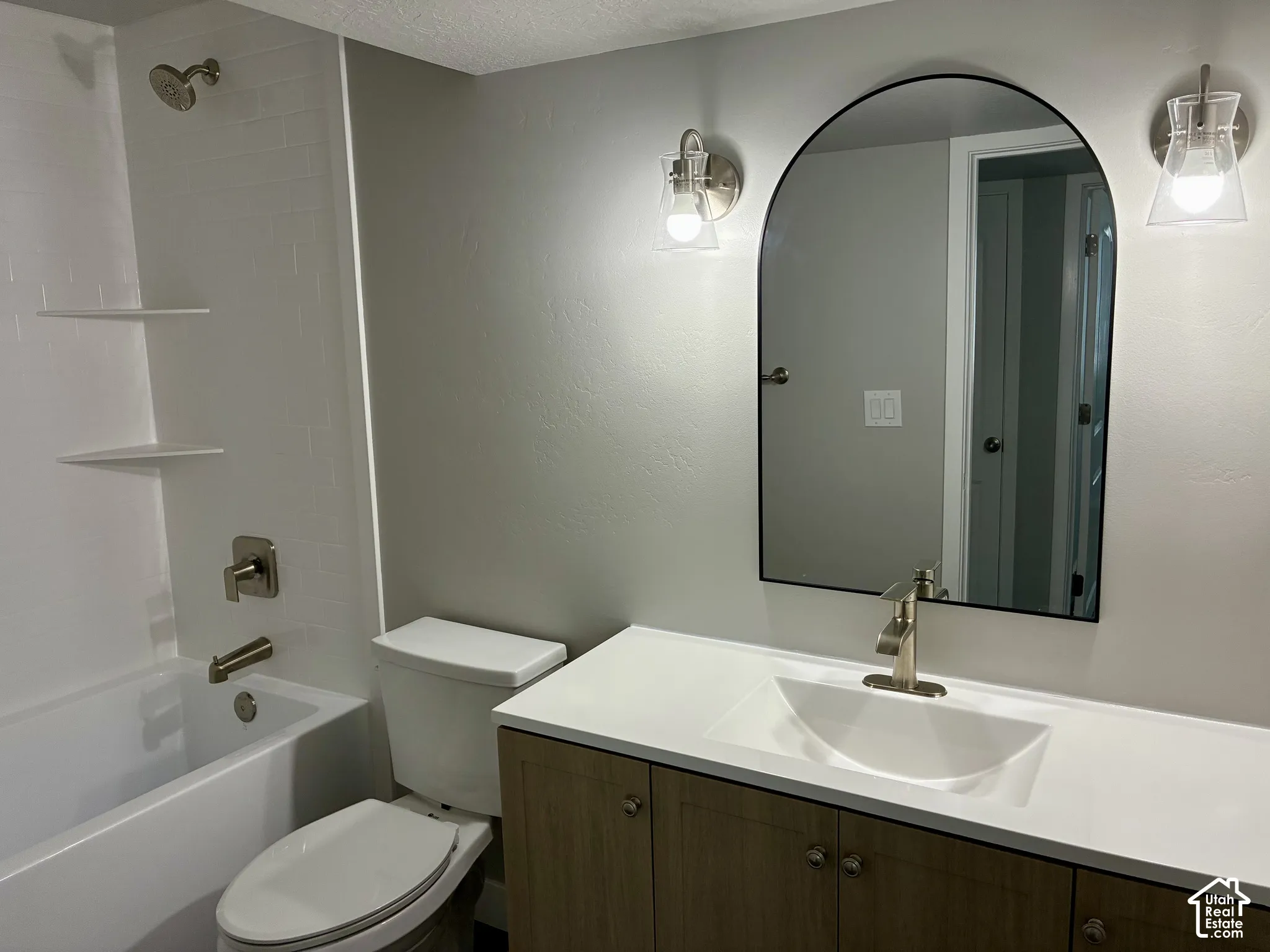 Vanity with wood-finish cabinetry and white countertop, undermount sink, arched mirror, brushed nickel faucet, and wall sconces
