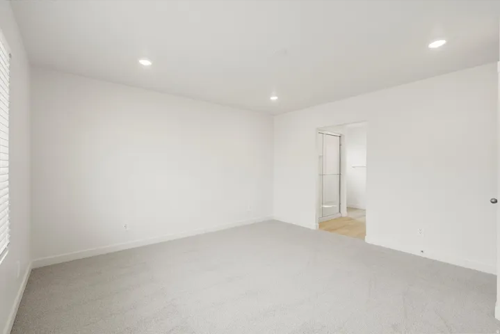 Spacious room featuring light gray carpeting, recessed ceiling lighting, a window with horizontal blinds, and a doorway opening to an en-suite bathroom with a glass-enclosed shower