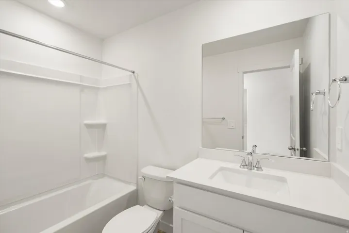 Bathroom featuring a white integrated tub and shower unit, a rectangular vanity mirror, a white vanity with an integrated sink, a chrome two-handle faucet, and a white toilet