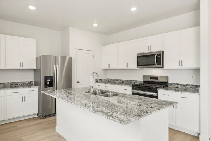 Bright kitchen featuring a central island with a double basin sink, white cabinetry with silver hardware, granite countertops, and stainless steel appliances including a refrigerator, microwave, and gas range