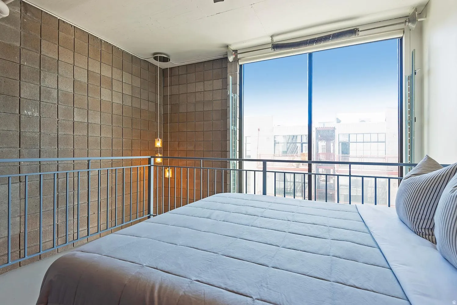 Spacious loft area featuring a full-height window, exposed concrete block wall, metal railing, and suspended pendant lighting