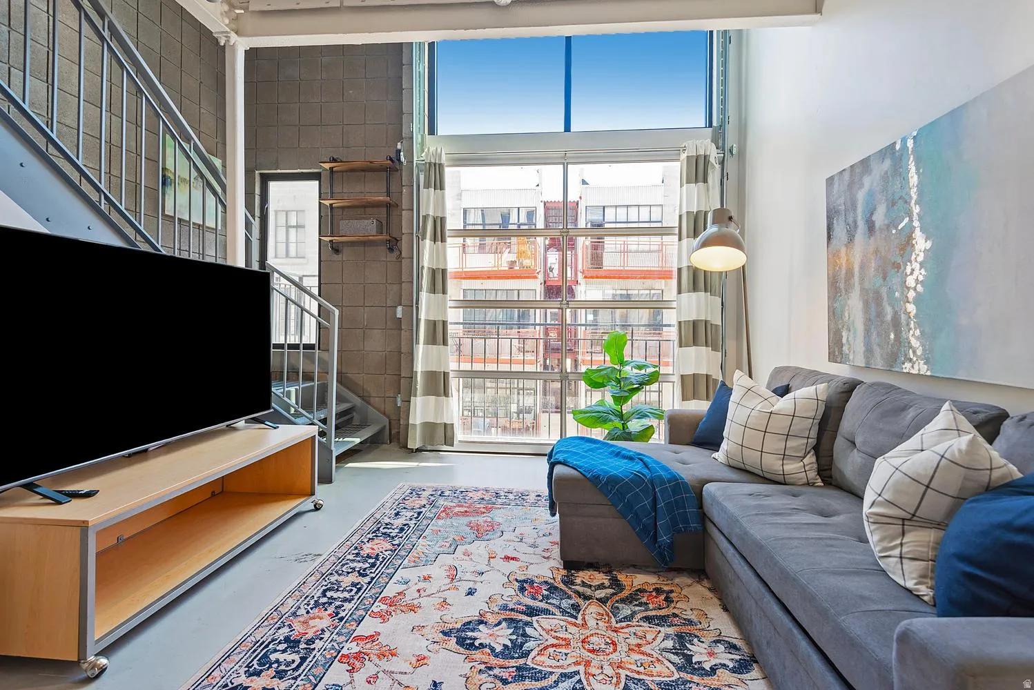 Spacious living area featuring high ceilings, concrete flooring, a large window wall, a gray sectional, and industrial-style stairs