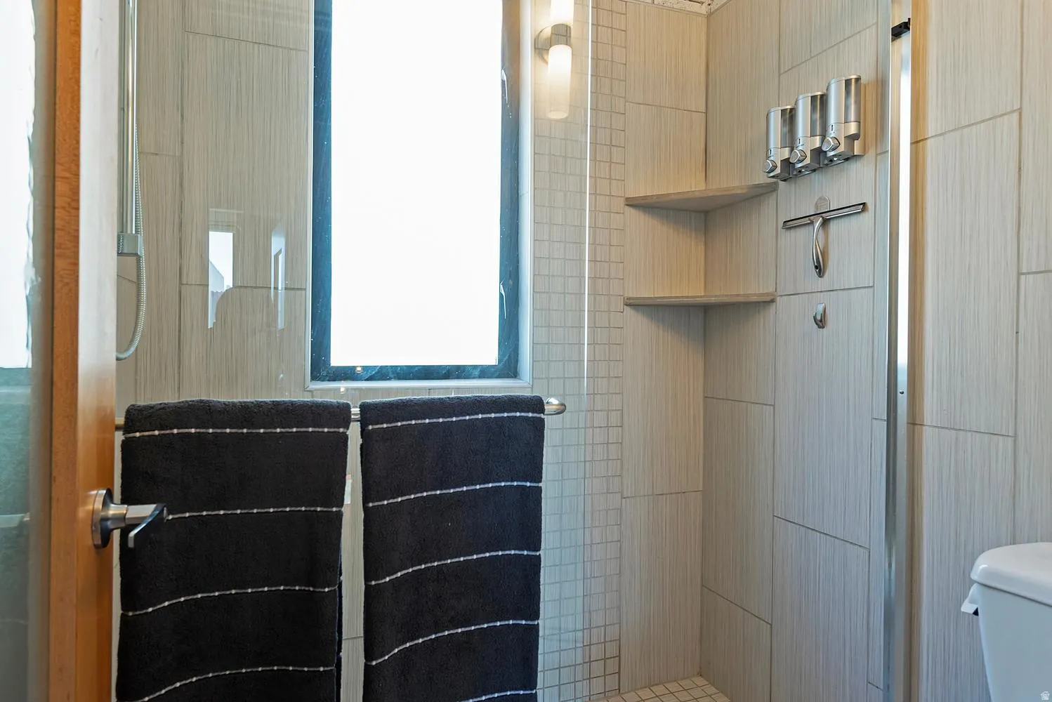 Shower enclosure featuring light-toned tile walls, built-in corner shelving, a recessed window, a wall-mounted fixture, and a glass partition