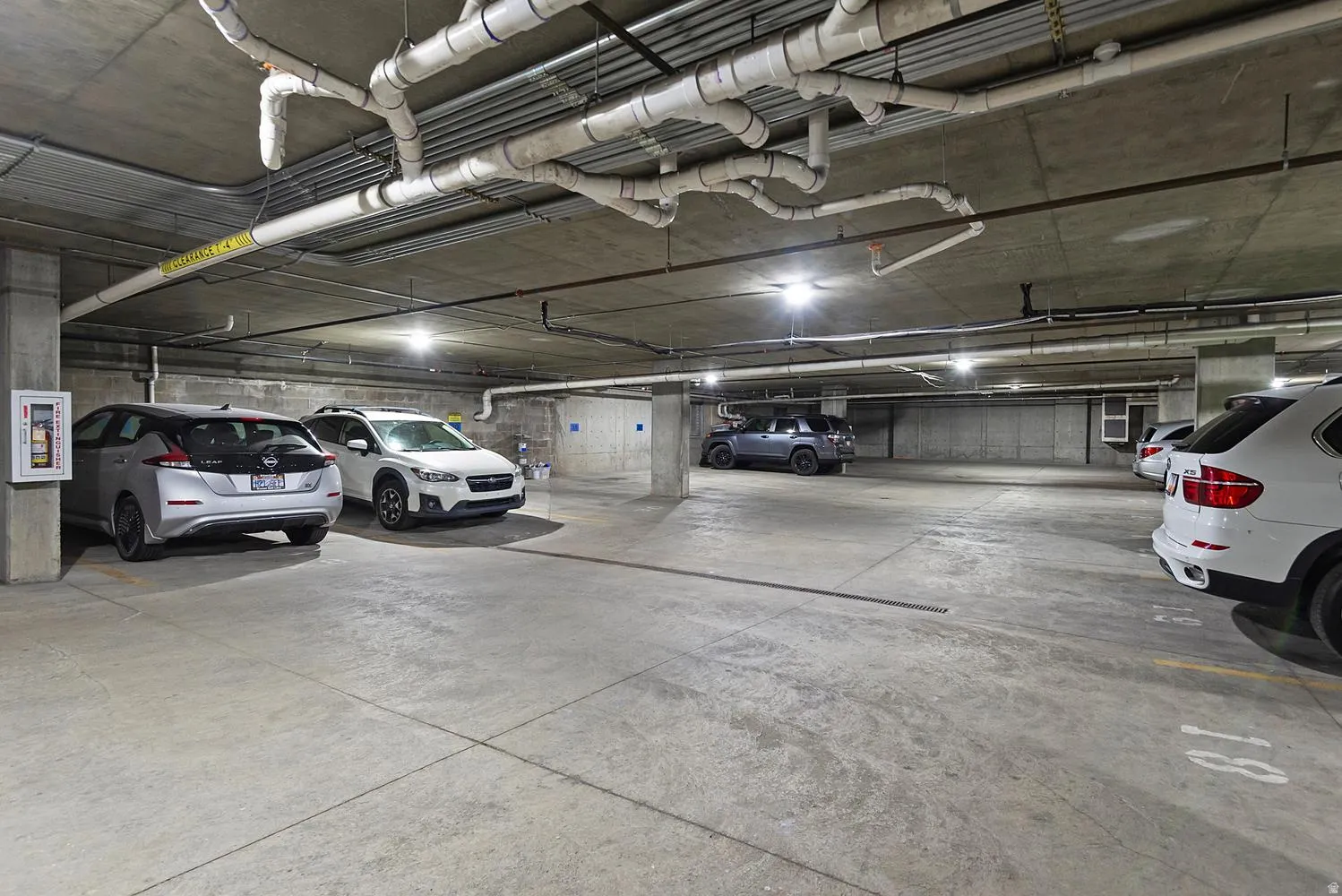 Underground parking facility with concrete flooring and ceiling, featuring exposed overhead piping and integrated lighting
