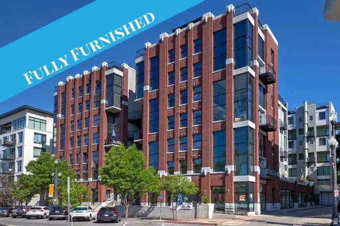 Modern multi-story building featuring extensive brickwork, large pane windows, multiple balconies with metal railings, and ground-level retail spaces